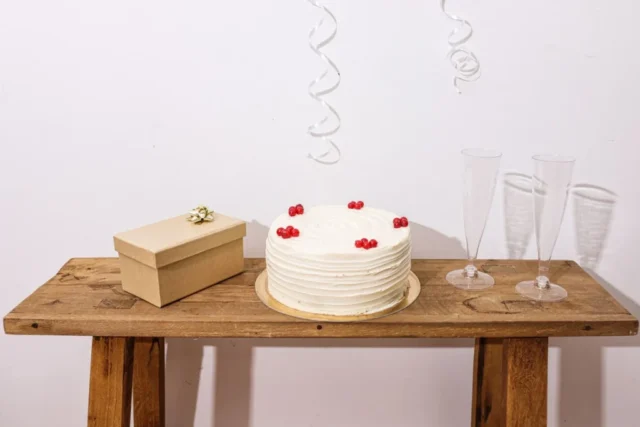 A white celebration cake, closed gift box and two glasses on a wooden table.