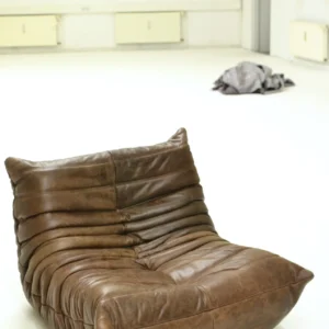 Low brown leather lounge chair with deep folds on a pale studio floor