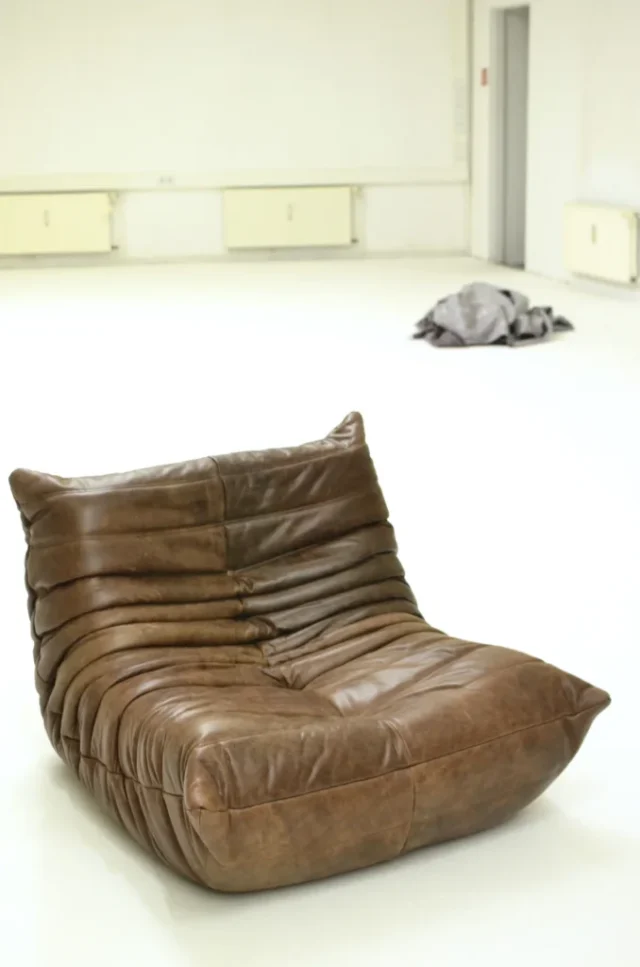 Low brown leather lounge chair with deep folds on a pale studio floor
